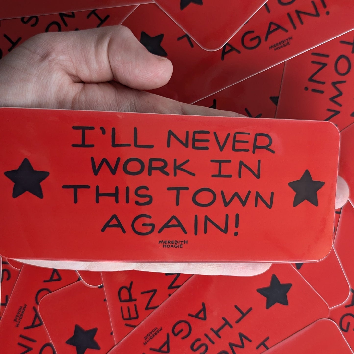 Red sticker with 'I'll Never Work in This Town Again!' text held by a hand against a background of similar stickers by Meredith Hoagie