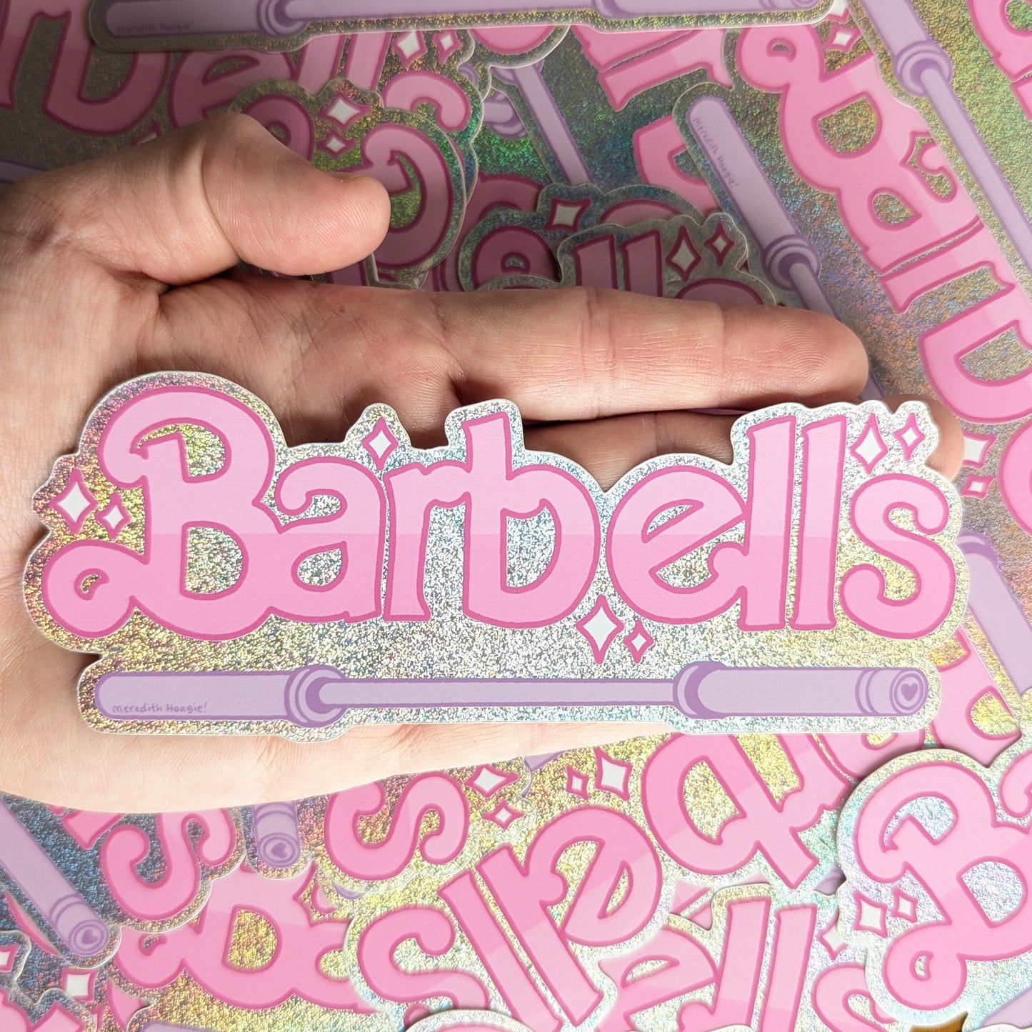 Pink holographic 'Barbells' sticker by Meredith Hoagie held over a background of similar stickers.