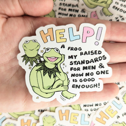 Sticker with a frog character and text 'HELP! A frog my raised standards for men & now no one is good enough!' created by Meredith Hoagie held by a hand.