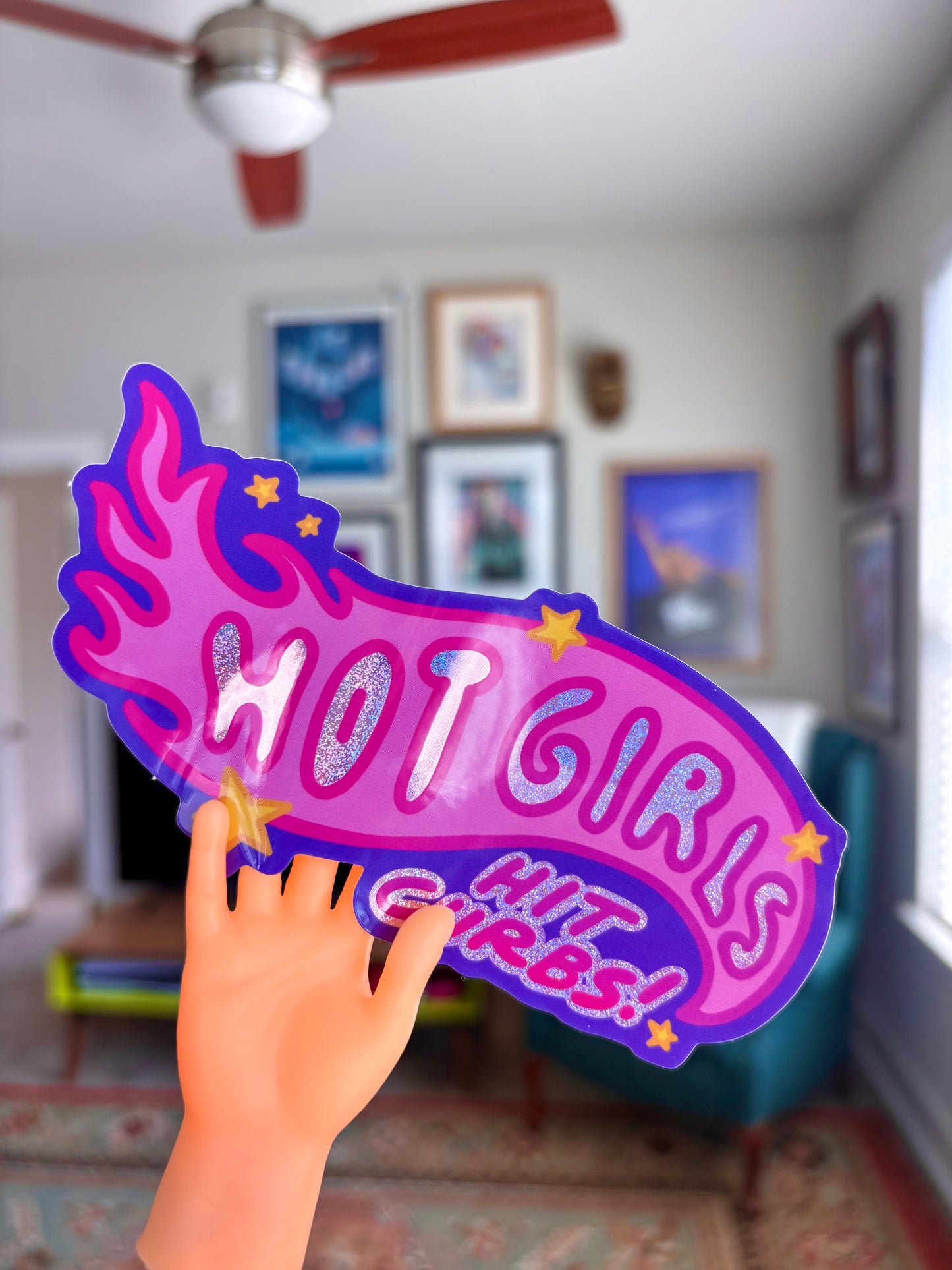 Plastic hand holding a 'HOT GIRLS HIT GIRLS' sign in a room with framed horror movie posters on the wall.
