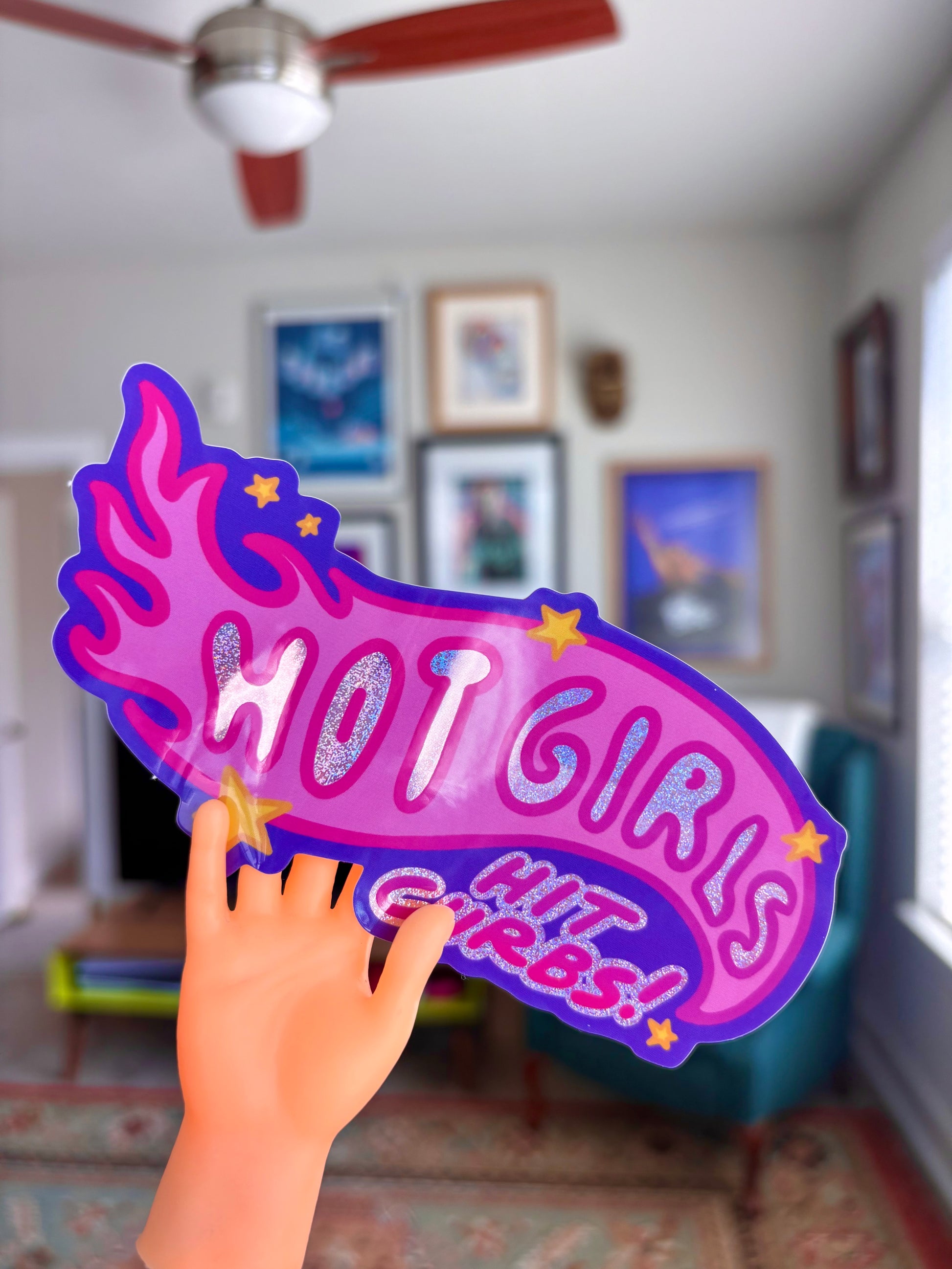 Plastic hand holding a 'HOT GIRLS HIT GIRLS' sign in a room with framed horror movie posters on the wall.