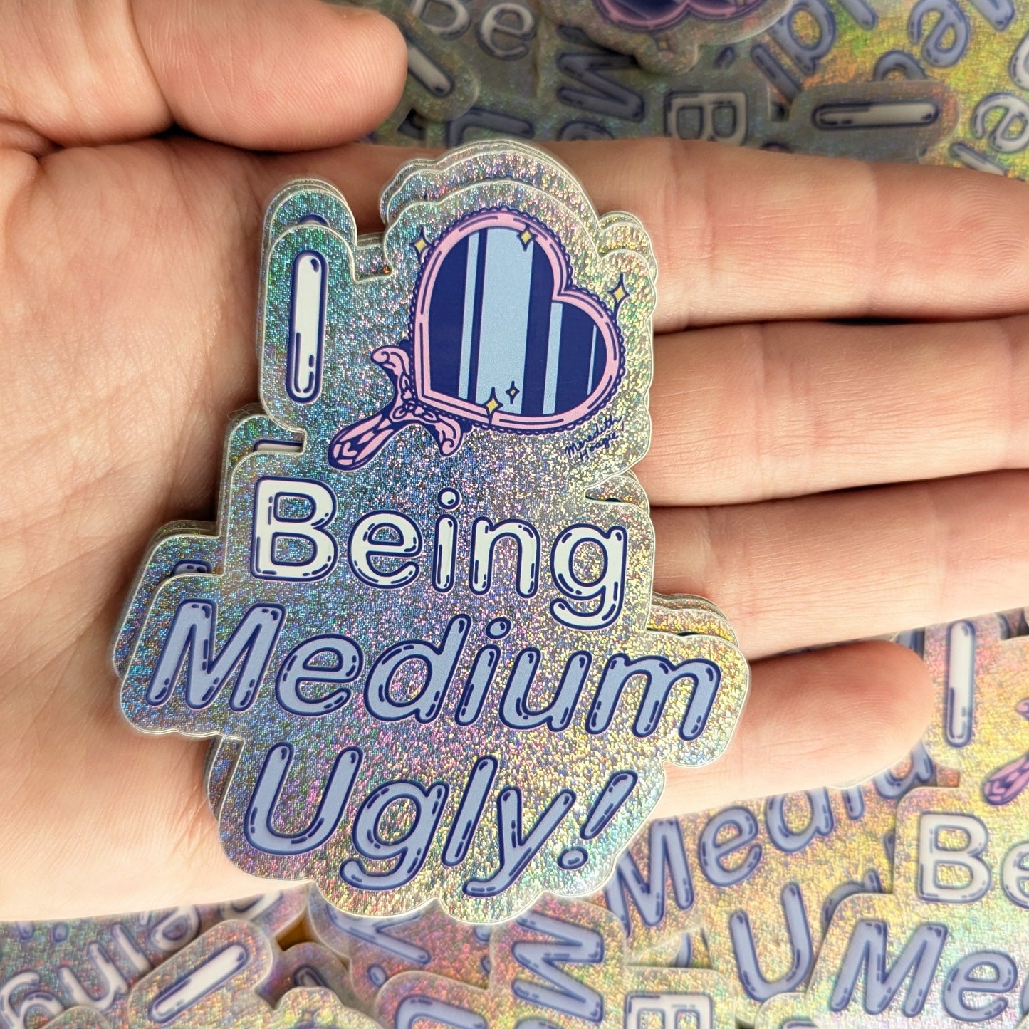Holographic sticker with text 'I Love Being Medium Ugly!' by Meredith Hoagie held by a hand