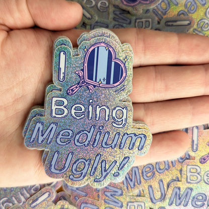Holographic sticker with text 'I Love Being Medium Ugly!' by Meredith Hoagie held by a hand
