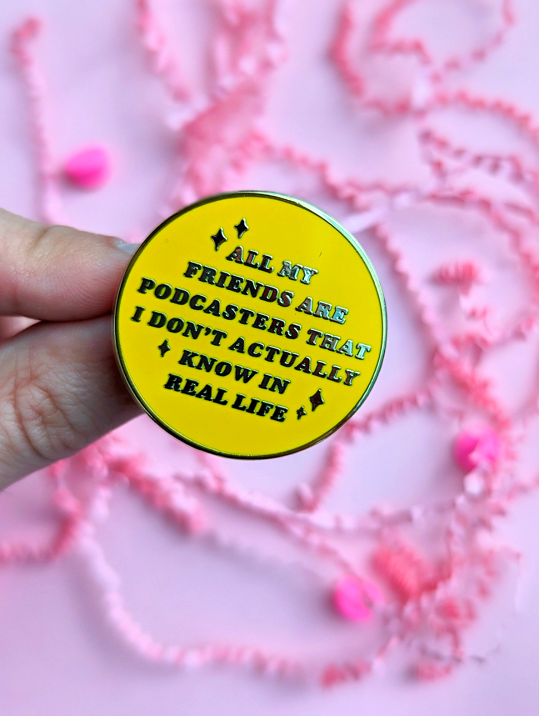 Cute Yellow Enamel Pin that says "All My Friends Are Podcasters That I Don't Actually Know In Real Life" held by a hand on a pink background | Ideal of the chronically online