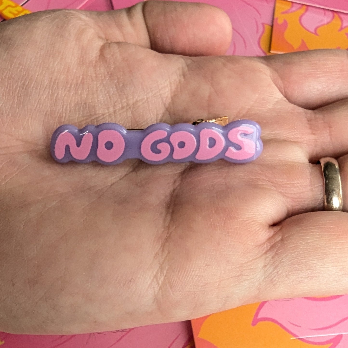 Hand holding a pink and purple 'No Gods' hair clip by Meredith Hoagie with cards in the background