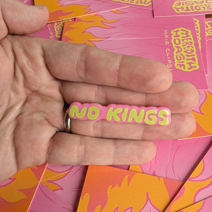 Hand holding a 'NO KINGS' hair clip by Meredith Hoagie with pink and yellow cards in the background